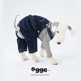 Dog wearing dark denim overalls over a striped shirt.