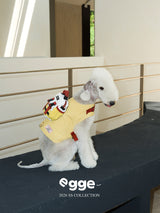 White dog wearing a yellow harness vest and backpack indoors.