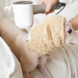 Dog wearing a beige plush ear-covering hood beside a hair dryer